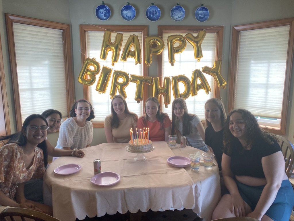 Eight girls sitting around a birthday cake at a pink table with gold happy birthday balloons behind them.