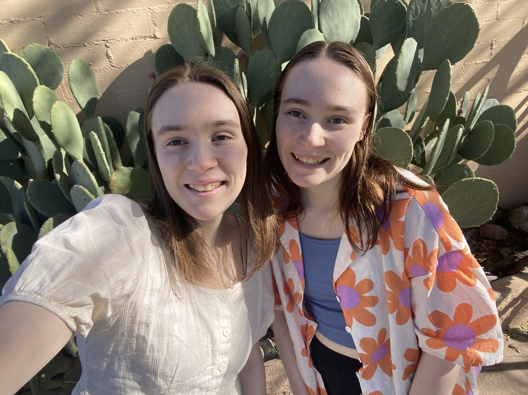 A selfie of two girls in front of a cactus, one in a white top and one in an orange flower top.