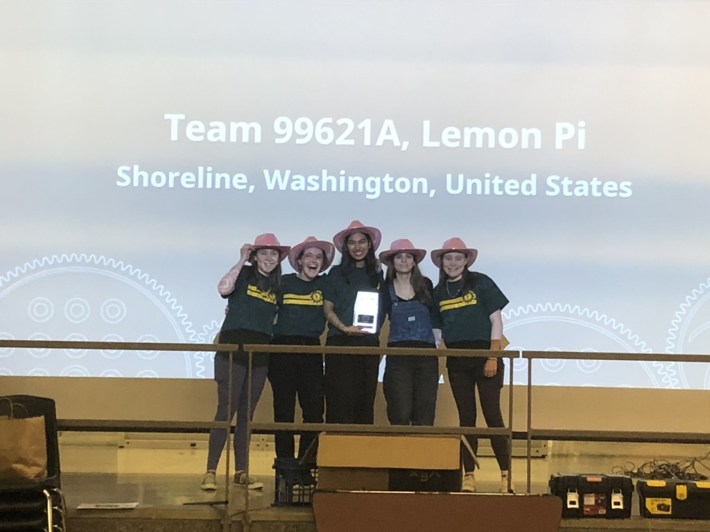 Five girls in pink cowboy hats standing in front of a screen with a trophy.
