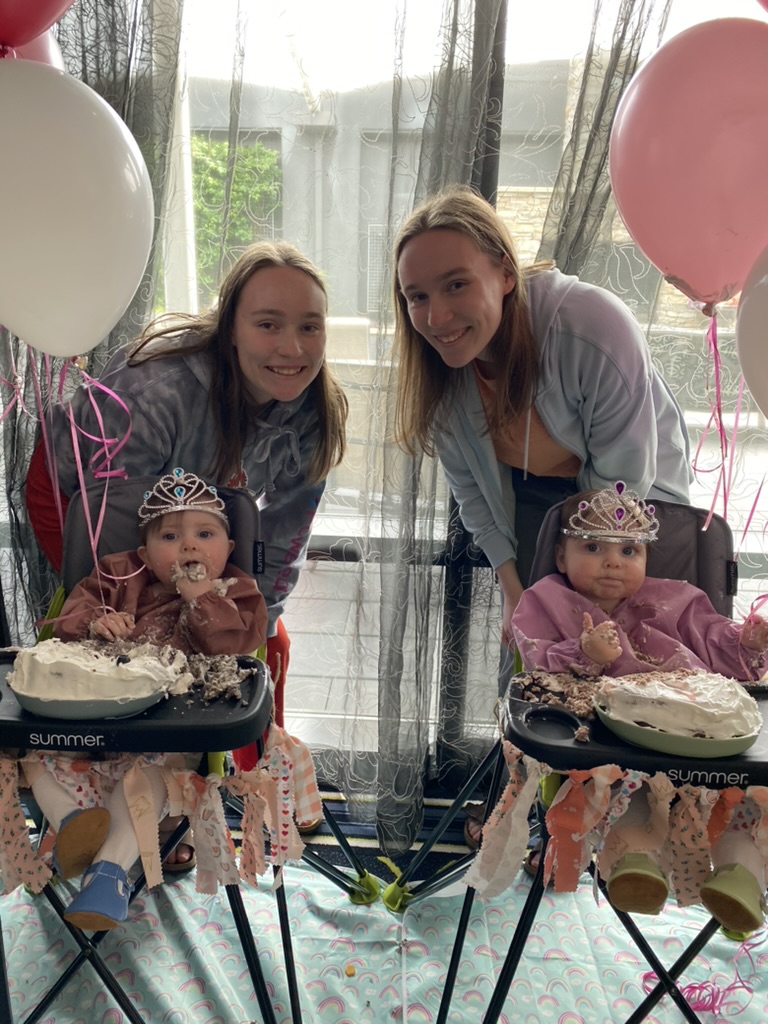 Twins smiling with baby twins who are eating cake in tiaras.