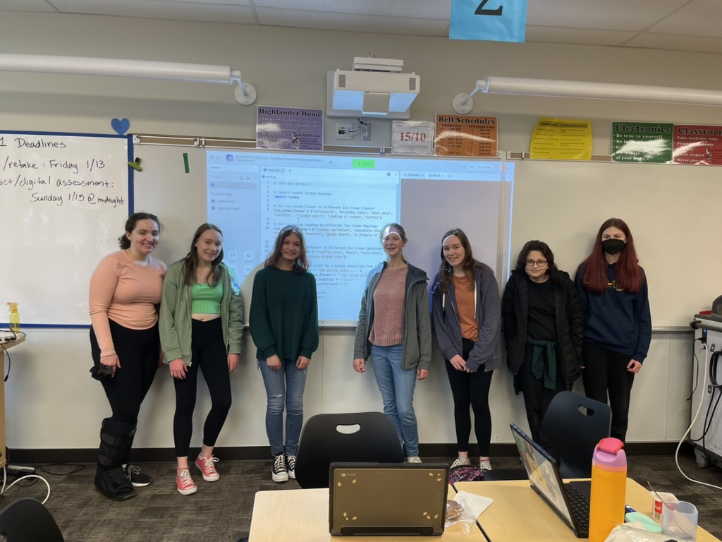 Seven girls standing in front of a Python random ice cream order generator program projected on a whiteboard in a classroom.