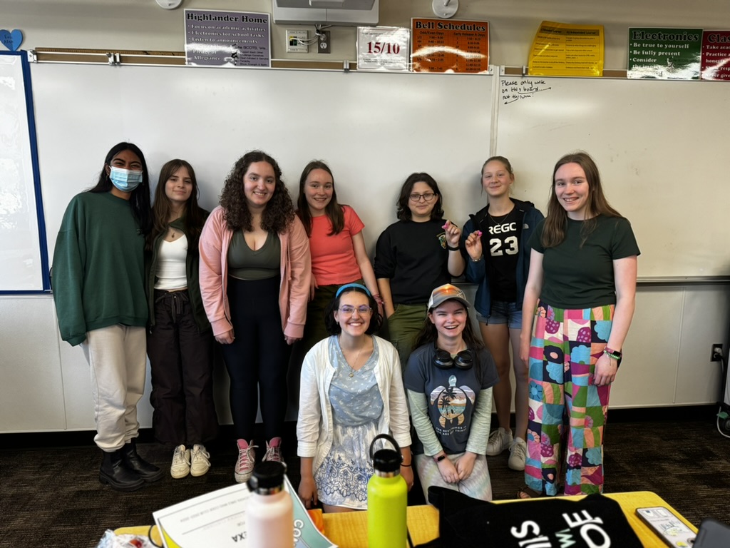 Nine girls smiling in front of a white board in a classroom. Some are holding pink 3D printed ducks.