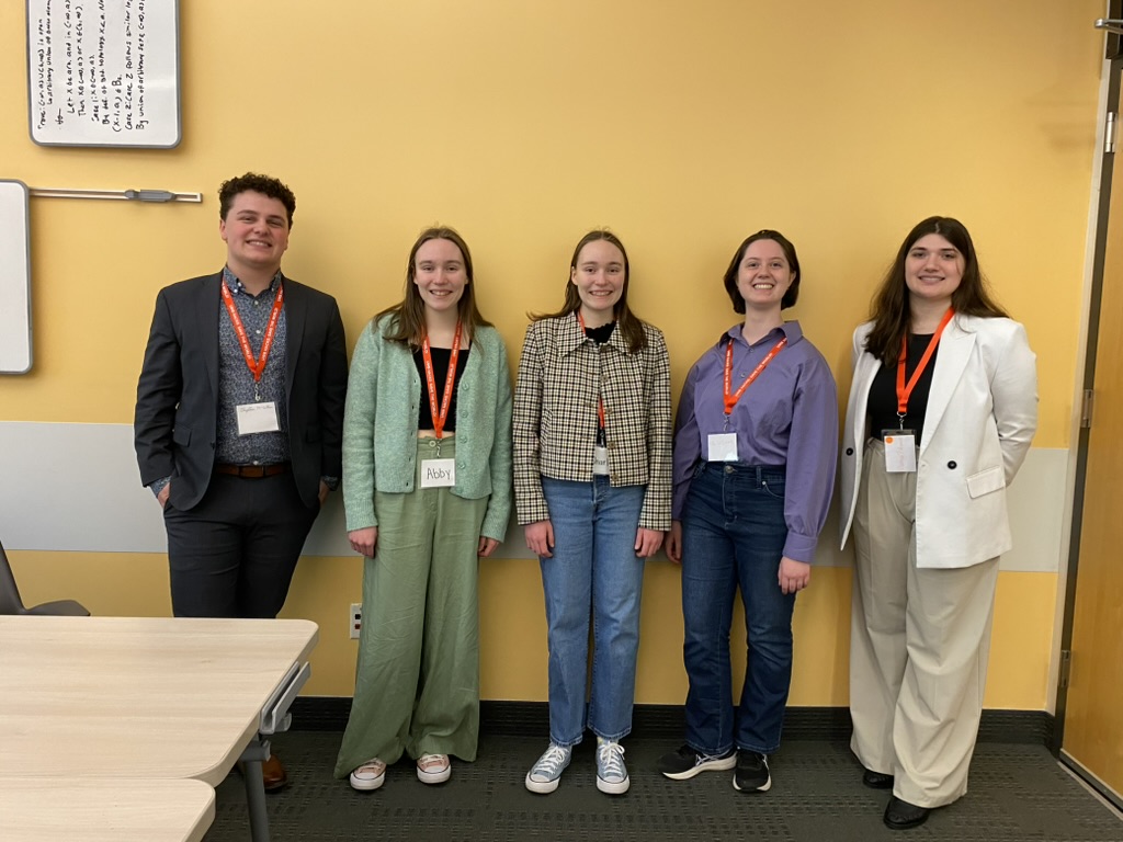 Five people dressed up in front of a bright yellow wall ready to pitch their project at a hackathon event.