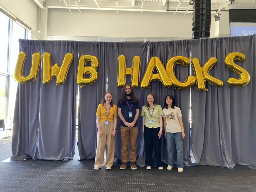 Four people standing in front of large golden balloons that read UWB HACKS.