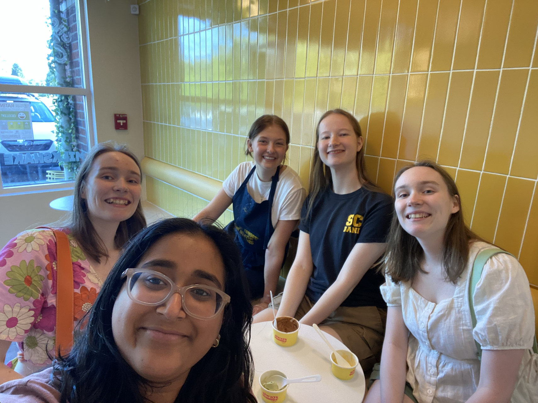 Five girls in front of a yellow tiled wall smiling with cups of ice cream.