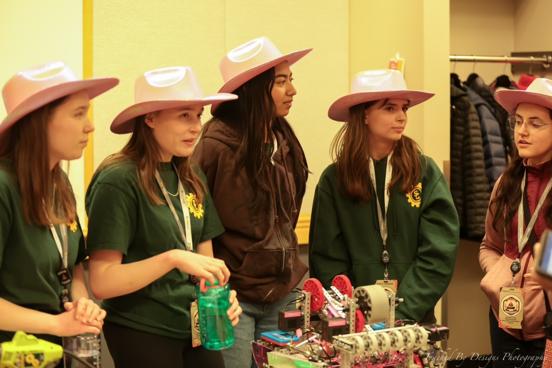 Five girls in pink cowboy hats being interviewed at a robotics competiton. Their robot is on the table in front of them.