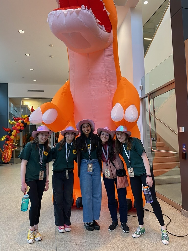 Five girls in pink cowboy hats in front of a giant orange inflatable dinosaur.
