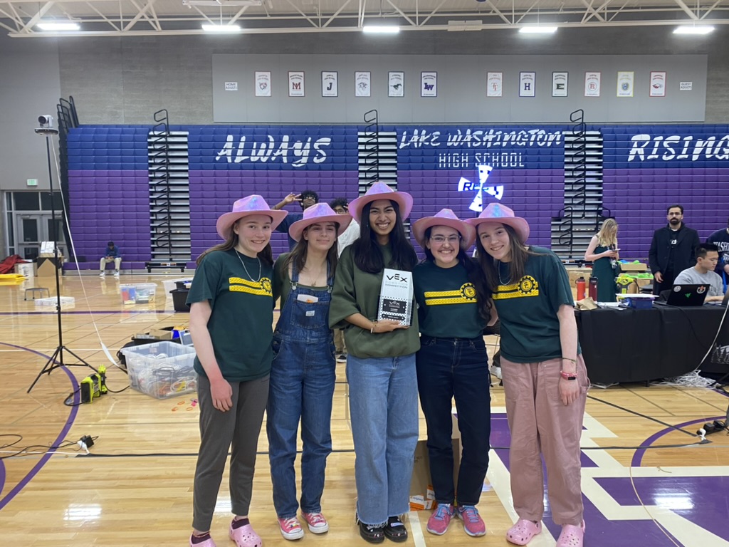 Five girls in pink cowboy hats holding a trophy in front of purple bleachers.