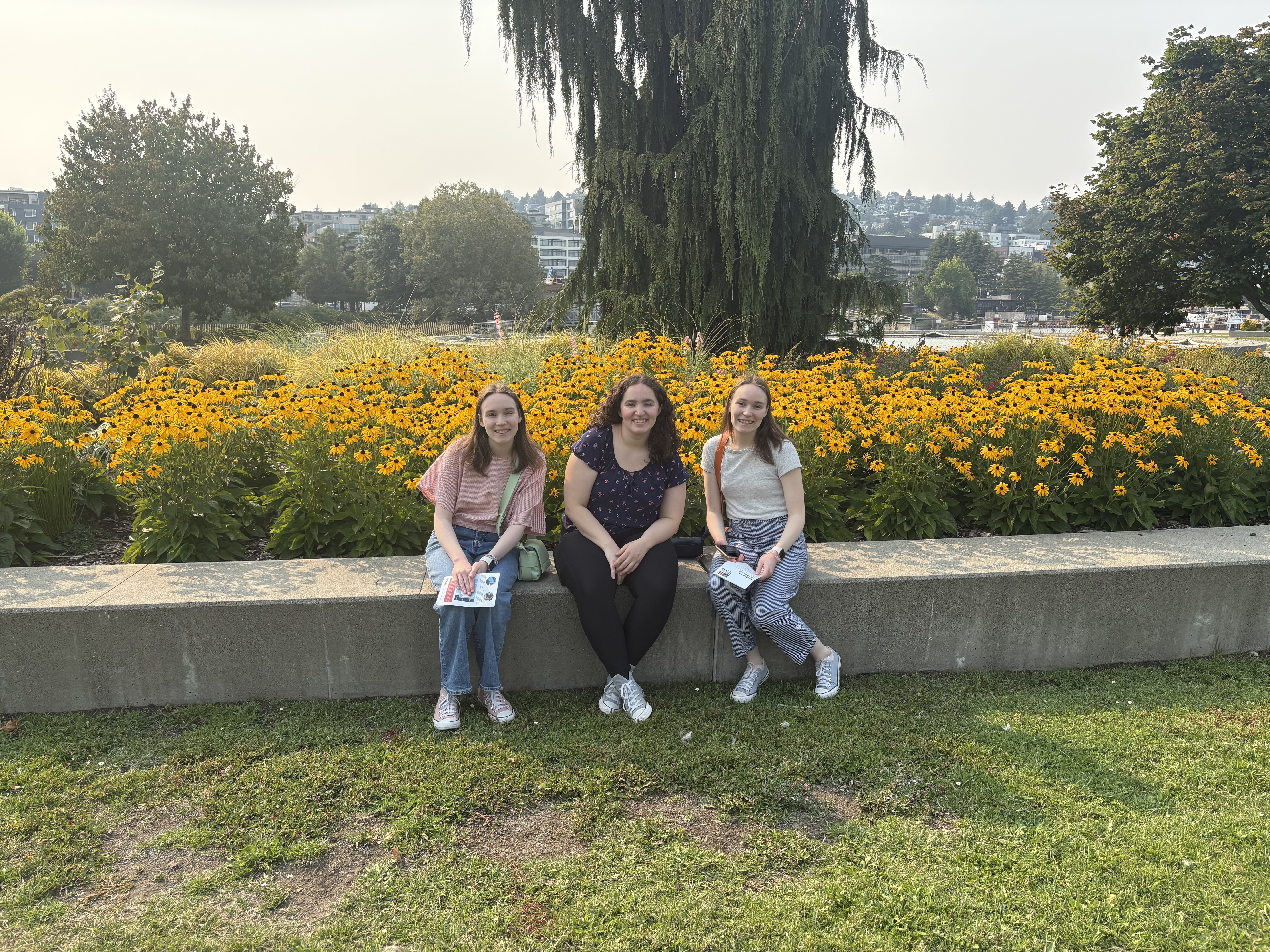 Three girls smiling on a bench in front of yellow flowers and trees.