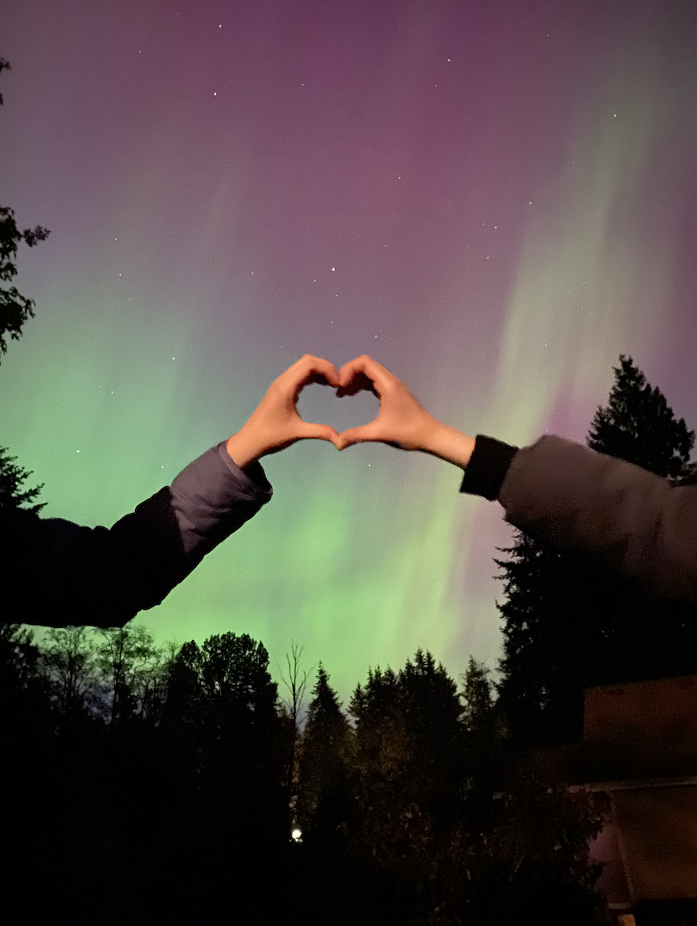 Two people making a heart with their hands in front of the northern lights.