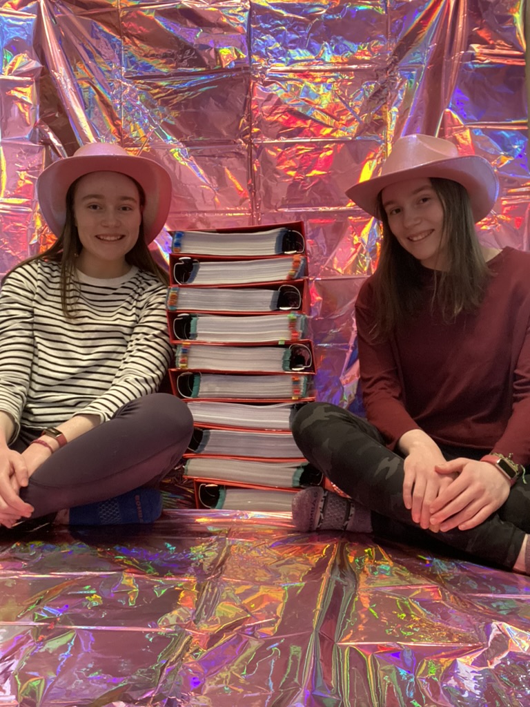 Two girls in pink cowboy hats sitting next to ten red binders stacked on top of each other.