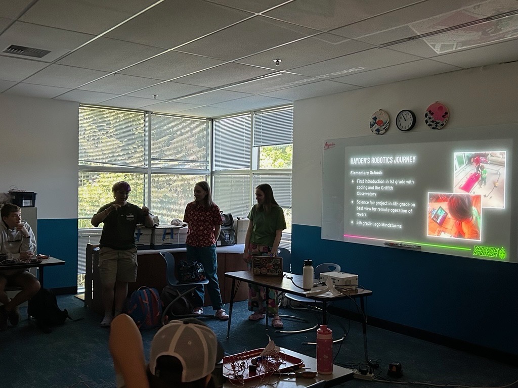 Three college students presenting in a school classroom. The room is dark but sun comes in through the windows.