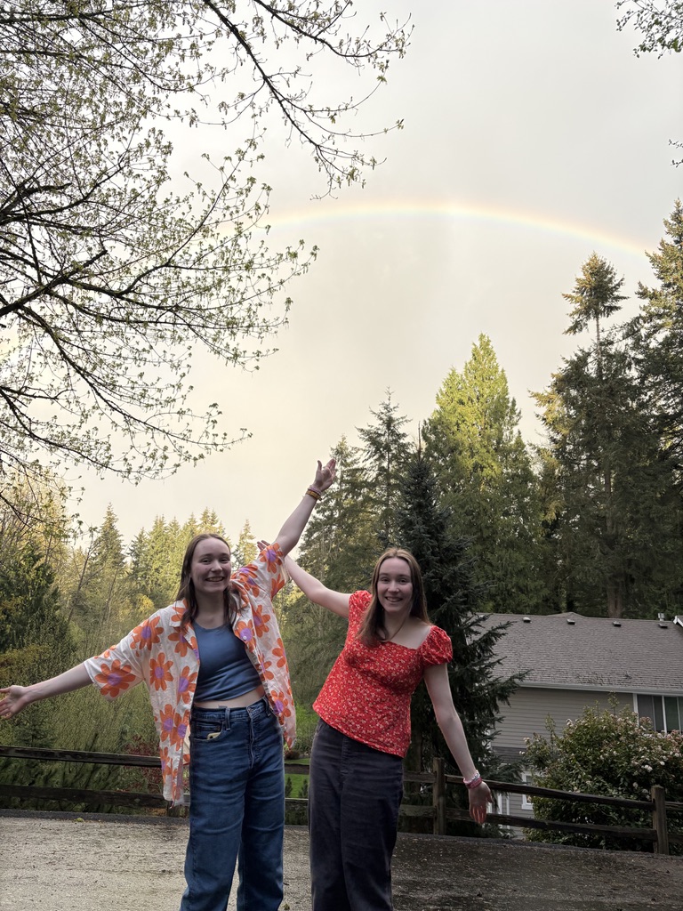 Two girls with their hands in the air in front of trees with a rainbow above them.