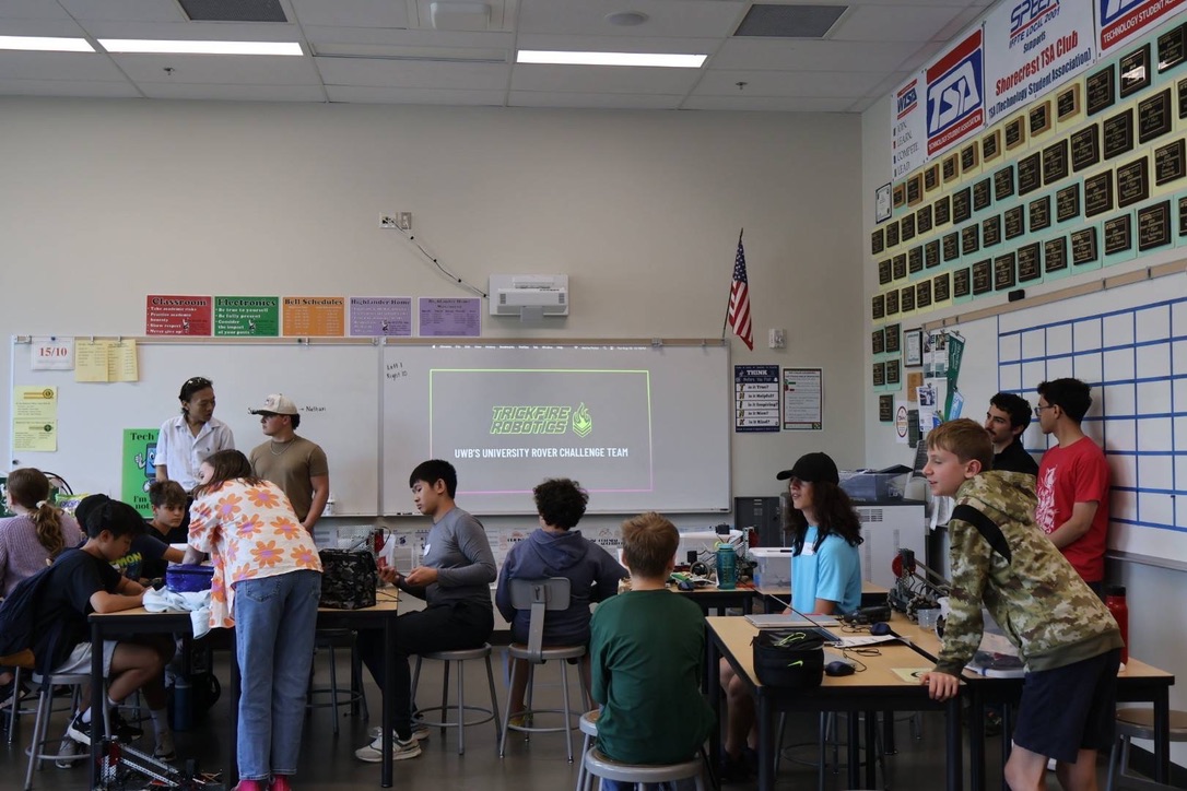 A classroom with many middle school summer campers working on robots at tables, college students preparing a presentation to the side.
