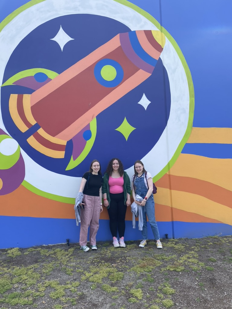 Three girls standing in front of a colorful rocket mural.
