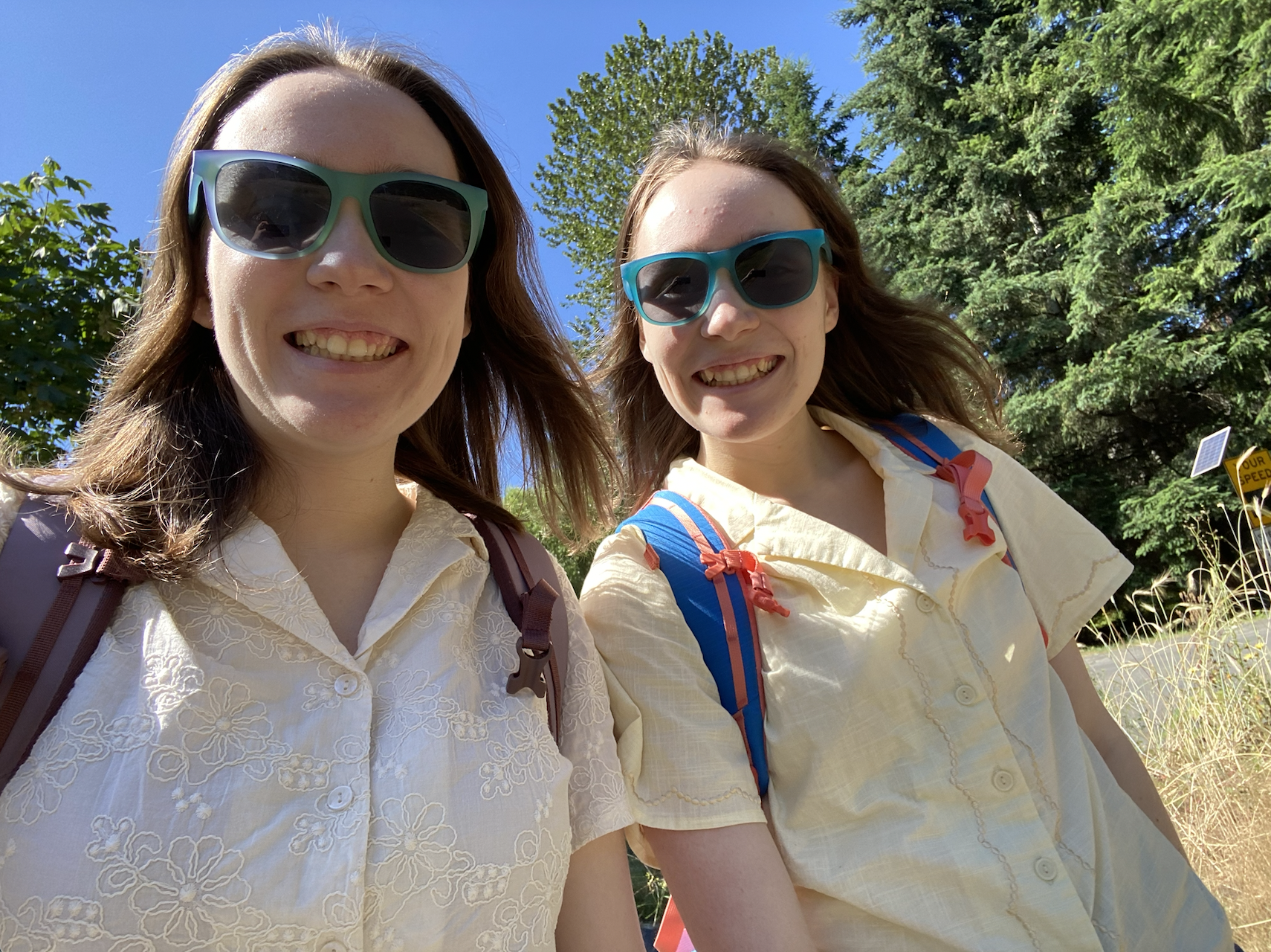 Two girls smiling in sunglasses with trees behind them.