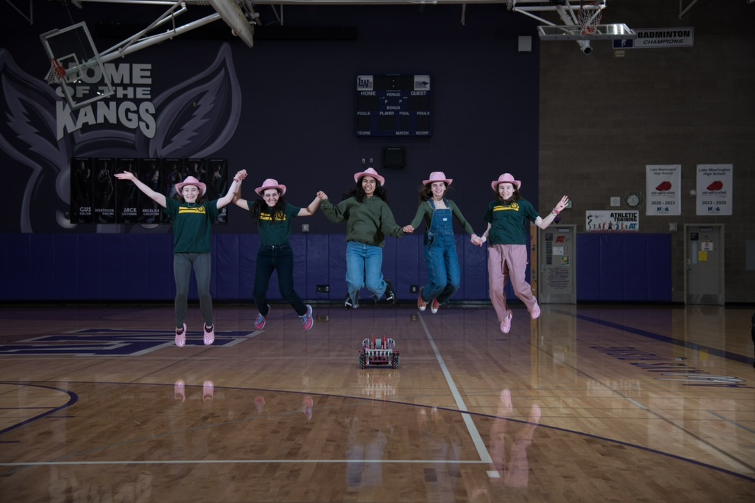 Five girls in pink cowboy hats holding hands and jumping in the air. Their pink and purple robto is on the floor in front of them.