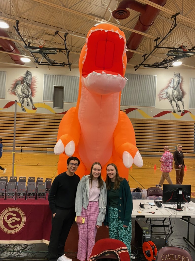 Three people smile in front of a giant orange inflatable dinosaur.
