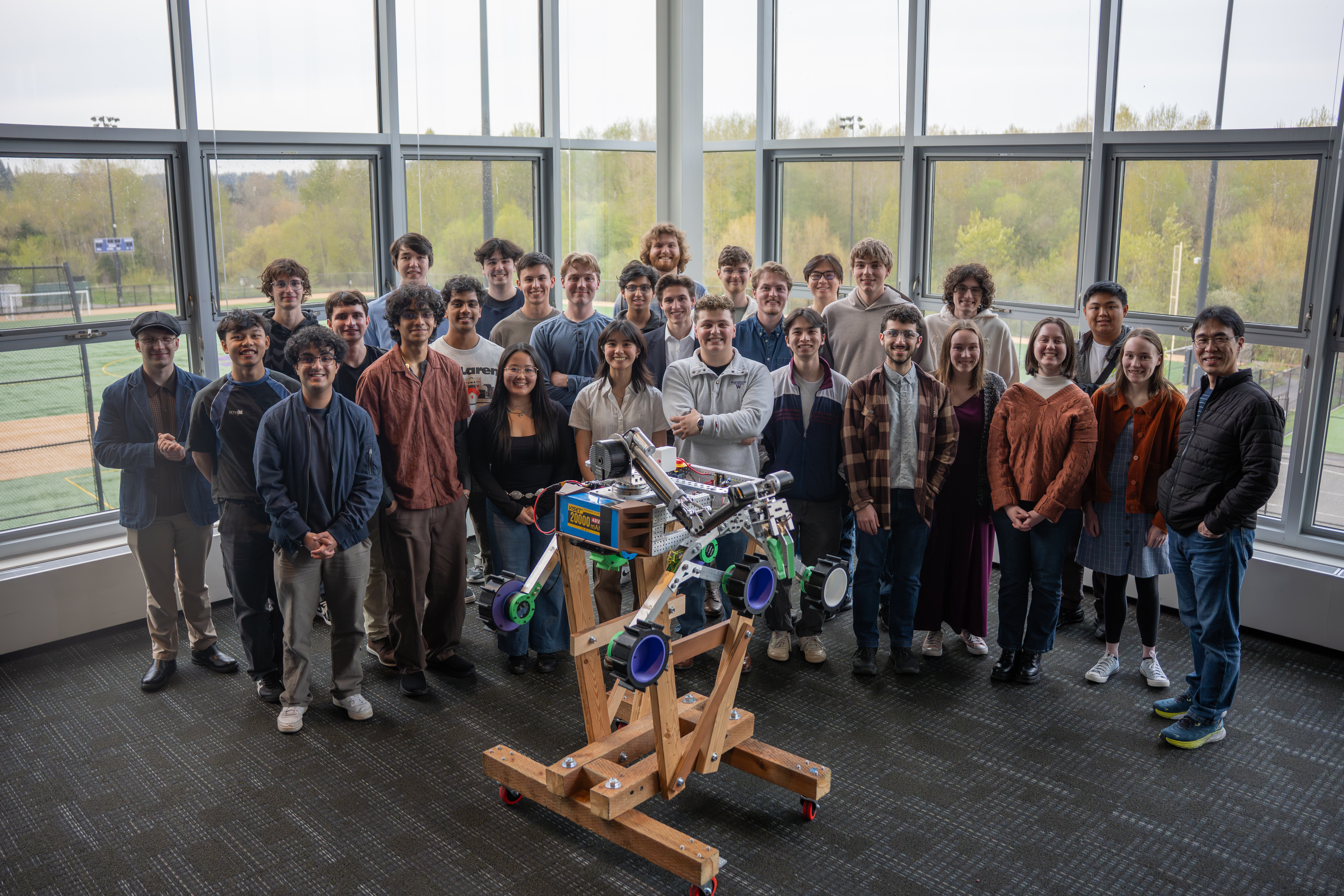 A group of students smile behind a gray rover on a wooden stand. Behind them are big windows.